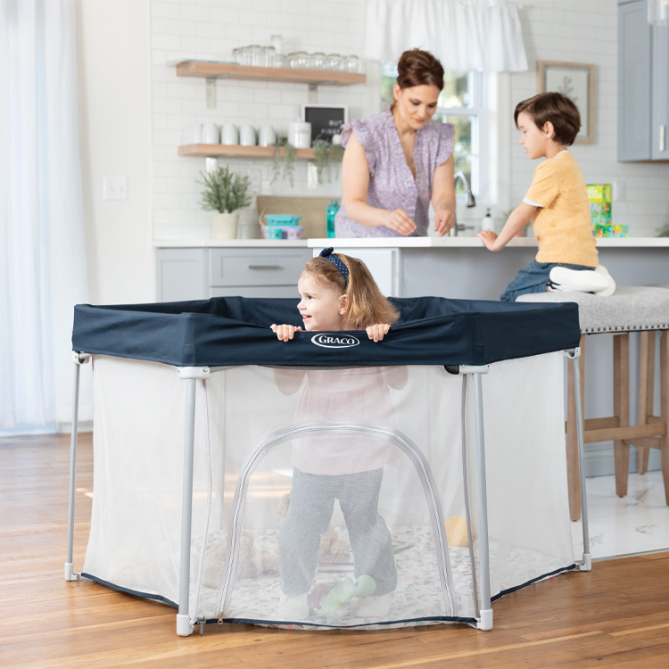 Kleinkind steht im Graco EverGo™ Laufgitter während die Mutter und der Bruder am Küchentresen stehen.