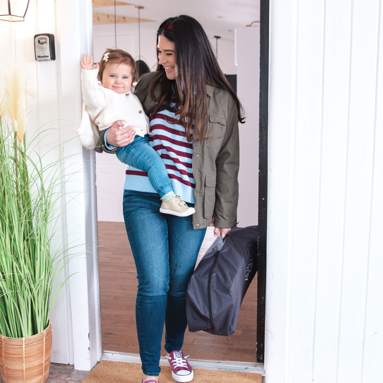 Mum holding toddler and Graco FoldLite Lightweight Travel Cot in doorway